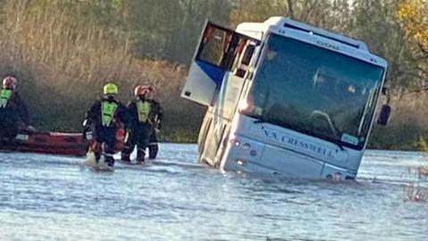 A bus tilting in flood water with rescuers approaching it pulling a boat