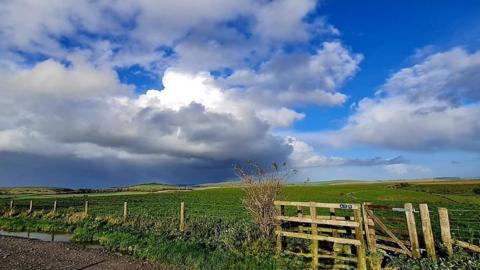 A huge swirl of grey/white cloud fills the two thirds of the sky from the left hand side with some blue sky showing in the right side. Below are green fields and mountains on the horizon.