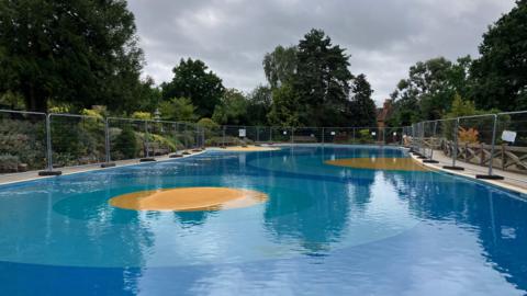 The paddling pool in Stoke Park, Guildford, Surrey, during a period of closure due to water contamination. Wire fencing can be seen around its parameters. 