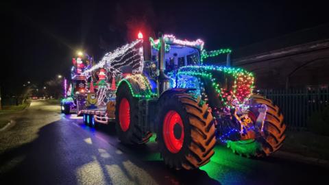 A tractor pulling a trailer, with both covered in lights and decorations. The photograph is taken at nighttime, and there are various different coloured lights covering the vehicle as well as inflatable Santas and Minions characters adorning it.
