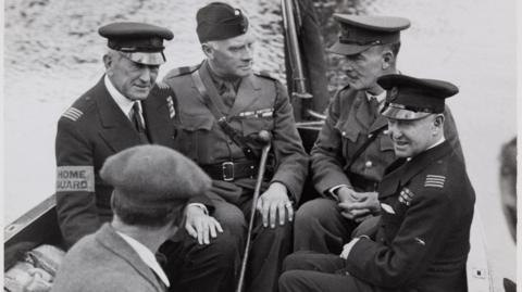 Five men, four wearing uniforms of the British Armed Forces sit in a small boat on the water. The fifth man in the black and white image has his back to the camera and is wearing a jacket with a white shirt collar visible and a flat cap.
