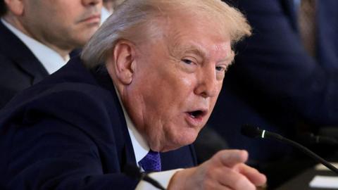 Donald Trump pointing towards the camera, sat next to other people whose faces are not in full view. He is wearing a purple tie and a blue jacket.