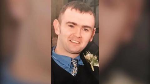 Damien Heagney with short brown hair, smiling while wearing a dark suit, blue shirt and patterned tie with a flower on it