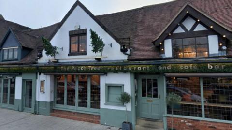 The front of a part timber-framed building with large windows and a low brown-tiled roof with the words The Peach Tree Restaurant running along the front on a green, leafy, sign