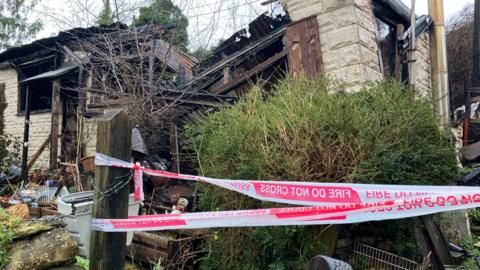 A burned down bungalow with fire service tape around it. The bungalow has caved in and the remains no are charred.