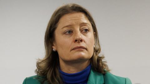 A woman with long brown hair. She is wearing a green blazer over a blue turtle neck jumper. She is also wearing silver earrings.