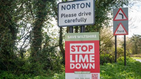 A village road sign for Norton with a temporary sign attached to the poles holding it up saying 'Stop Lime Down'. One the side of green verge with trees.