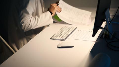 A doctor is sat in a white coat at a white desk with a computer, keyboard and documents. She is sifting through the paperwork. 