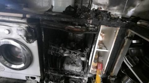 A dishwasher with fire damage . The are is blackened, part of the neighbouring dishwasher has melted.