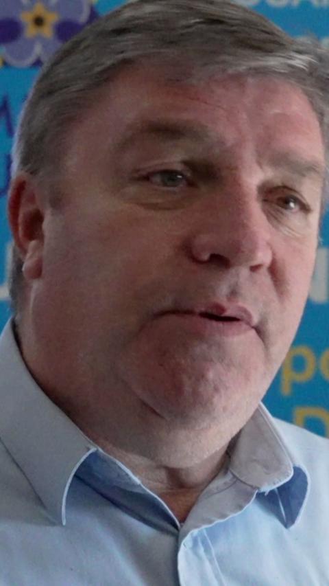 A man with grey hair and eyebrows, talking to the camera. He wears a blue shirt.