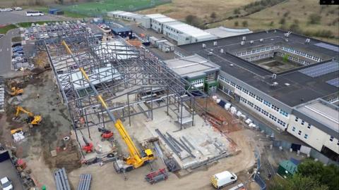 A large yellow crane is in the foreground. The metal structure of the new part of the school can be seen. On the right hand side is the rest of the school.