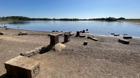 A reservoir which is very low, under blur skies. There are lots of stone blocks on the edge on the way into the water.
