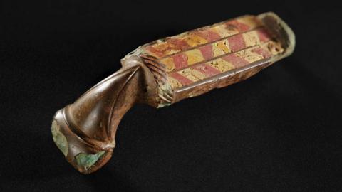 The brooch is a a rectangle shaped object with a brown taper shaped end. The rectangle shaped section is decorated with a red and yellow chevron-like pattern. The brooch has been photographed against a black background. 