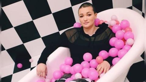 Caitlin posing in a bath (fully clothed) with pink balls all around her on a black and white background