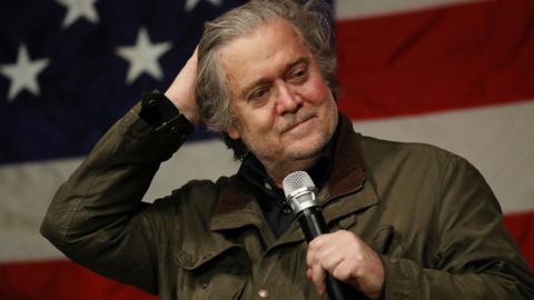 Steve Bannon at rally in front of US flag holding microphone