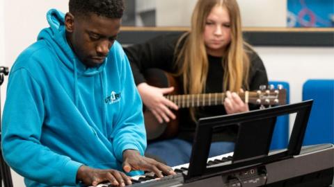 A teenage boy plays a keyboard, watched by a teenage girl playing guitar