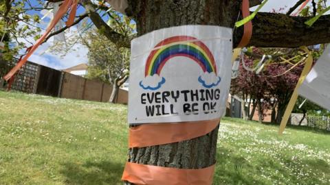 A sign pinned to a tree shows a rainbow and the message 'Everything will be ok'