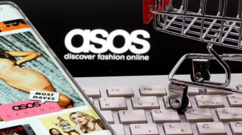 A smartphone with the Asos app, a keyboard and a shopping trolley are seen in front of a displayed Asos logo in an illustration picture