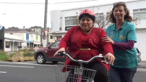 A woman rides a bike helped by a volunteer