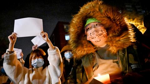 Protesters march along a street during a rally for the victims of a deadly fire as well as a protest against China's harsh Covid-19 restrictions in Beijing on November 28, 2022. Photo shows two people wearing facemasks, one holding a blank sheet of white paper and one with a candle.