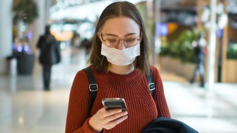 Stock image of a young woman wearing a face mask and holding a smartphone