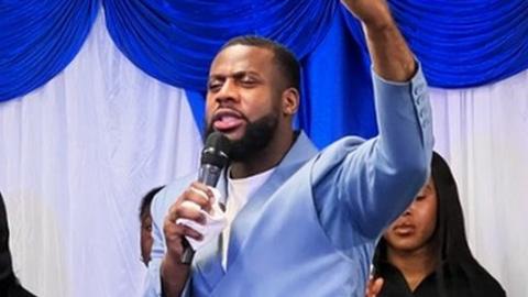 Pastor Onyeka standing on stage with a choir, speaking into a microphone with his eyes shut and hand raised