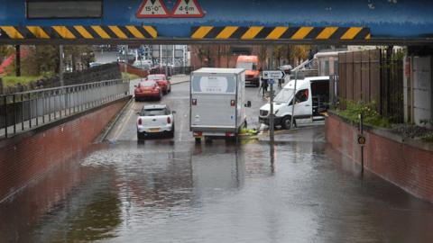 A road which passes under a rail bridge is flooded. Vehicles have been stopped by the water.