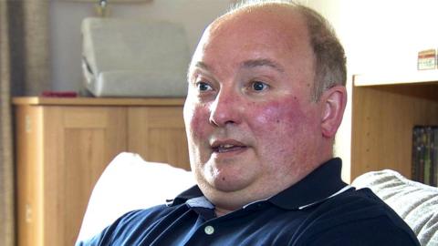 John Clarke sits at home in a blue shirt