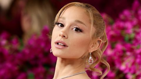 Ariana Grande, a woman looking at the camera with a tilted neck. She is wearing a necklace with the background blurred of pink flowers.