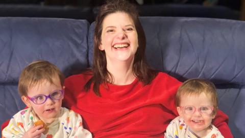 A white woman in a red jumper with brown hair smiles at the camera with her two toddler daughters sat either side of her, both girls wear colourful glasses