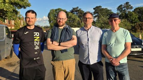 Four men stand side by side on a sunlit roadside, with parked vehicles and trees behind them.