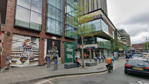 A Google Street View image shows the scene on Penn Street through Cabot Circus. On the pavement a person sits on a public bench in front of a shop window covered with an advert. High up on a building behind them to their left, a sign reads "Cabot Circus". 