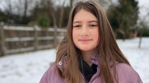 A child, standing outside, looking at the camera, with long straight dark hair, and a pink coat, with a dark top underneath. There is snow on the ground and behind her.