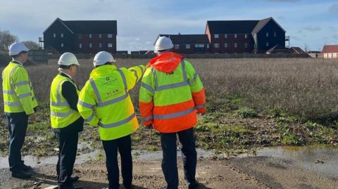 A group of council officials on the site of Wolsey Park in Rayleigh. There are three men dressed in hi-vis jackets and hard hats looking across an empty field towards houses in the distance.