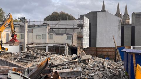 A Bulldozer demolishes old buildings in Truro , piles of rubble and are all around. The spires of Truro Cathedral can be seen in the background.