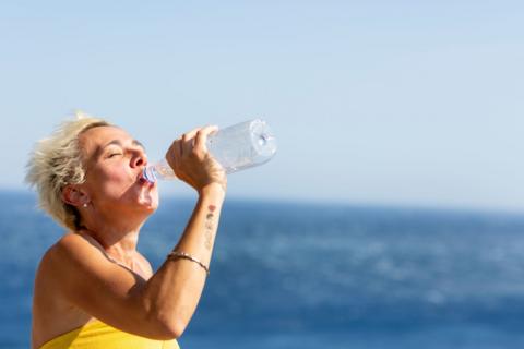 A woman with short blonde hair drinks from a plastic water bottle by the sea.