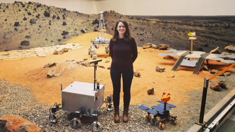 A woman wearing a brown jumper and black trousers stands on a simulated surface of Mars with orange sand and stones, with prototypes at her feet