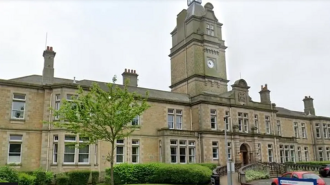 A long large Victorian building takes up most of the image with steps leading up to the entrance and a ramp to one side. A large clock tower sits in the centre of the building and there are several chimneys