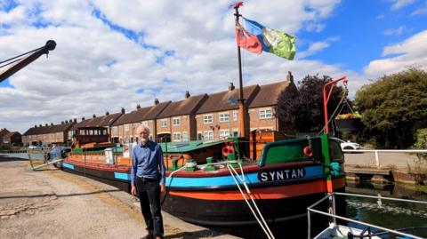 Iain Campbell stands next to a moored barge called Syntan. A Yorkshire flag flies from the mast alongside a Red Ensign. It is moored along a canal (Beverley Beck). The sun is shining and there a few clouds in the bright blue sky above. There are some terraced houses in the background.