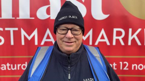 Allan Hartwell - a man wearing black, square framed glasses, a black beanie with 'Market Place' written in white letters, a dark zip jacket and a blue hi-vis jacket smiles at the camera. He is stood in front of large red sign about the Belfast Christmas Market.