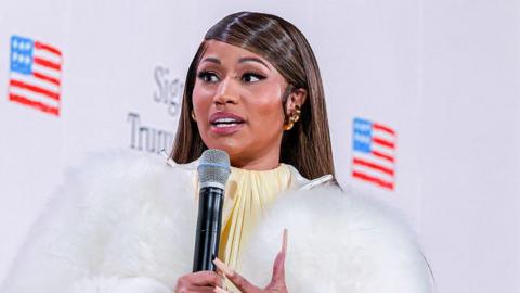 Nicki Minaj holds a microphone. She is wearing a white, fluffy coat.