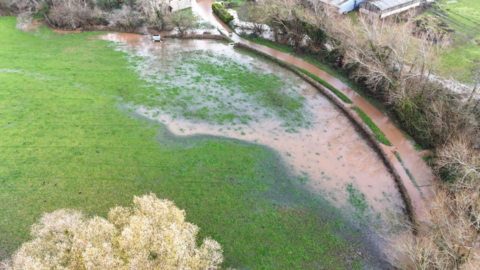 An aerial shot of a flooded field and country road. The water is brown and muddy and the road is lined by trees with no leaves. There are farm buildings at the top of the shot