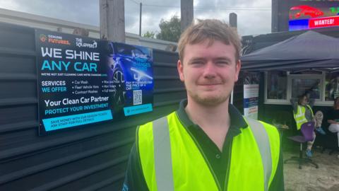 Luis Green stands outside his own car washing business in Bolton. Wearing a  dark polo shirt with a yellow hi-viz tabard, Luis is photographed in the scrapyard where his company is based.