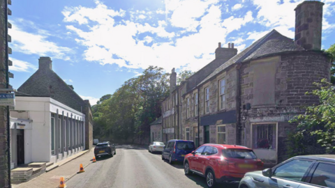 Union Street in Wick with cars parked on both sides of the road and some police no-waiting cones. There are brick houses down the right hand side and what looks like some kind of showroom on the left with trees in the distance.