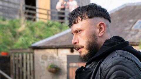 Richard Gadd stares intensely. He is wearing a black leather jacket and hoodie and is outside. Stone and slate buildings can be seen behind him. There are two people far behind on a terrace.