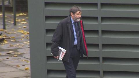 Alan Harris wearing a black coat with a red inner lining, dark suit, blue shirt and a dark blue tie with yellow markings on it walks past a green metal structure which has ventilation slots on the exterior. He is carrying a couple of notepads. Mr Harris has a greying beard and greying hair.