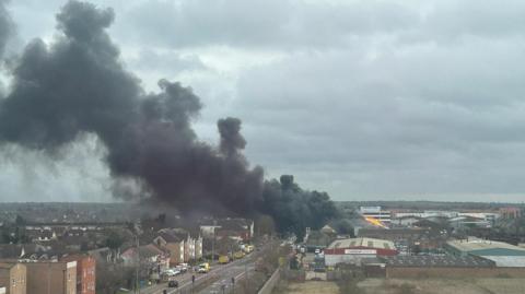 A huge plume of black smoke rises high into the air from a fire on an industrial estate. Flames are visible on the right of the image. There are industrial buildings on the right of the picture and residential properties on the left. 