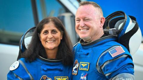 Sunita Williams and Butch Wilmore stand smiling for the camera while wearing astronaut suits