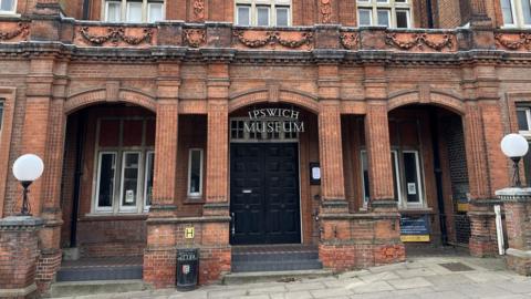 The entrance into Ipswich Museum. It is a red brick building with a large black door. The name of the museum hangs over the door.