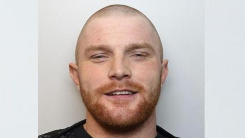 Darren Roberts' custody image. He has a short ginger beard, cropped hair and is smiling into the camera.